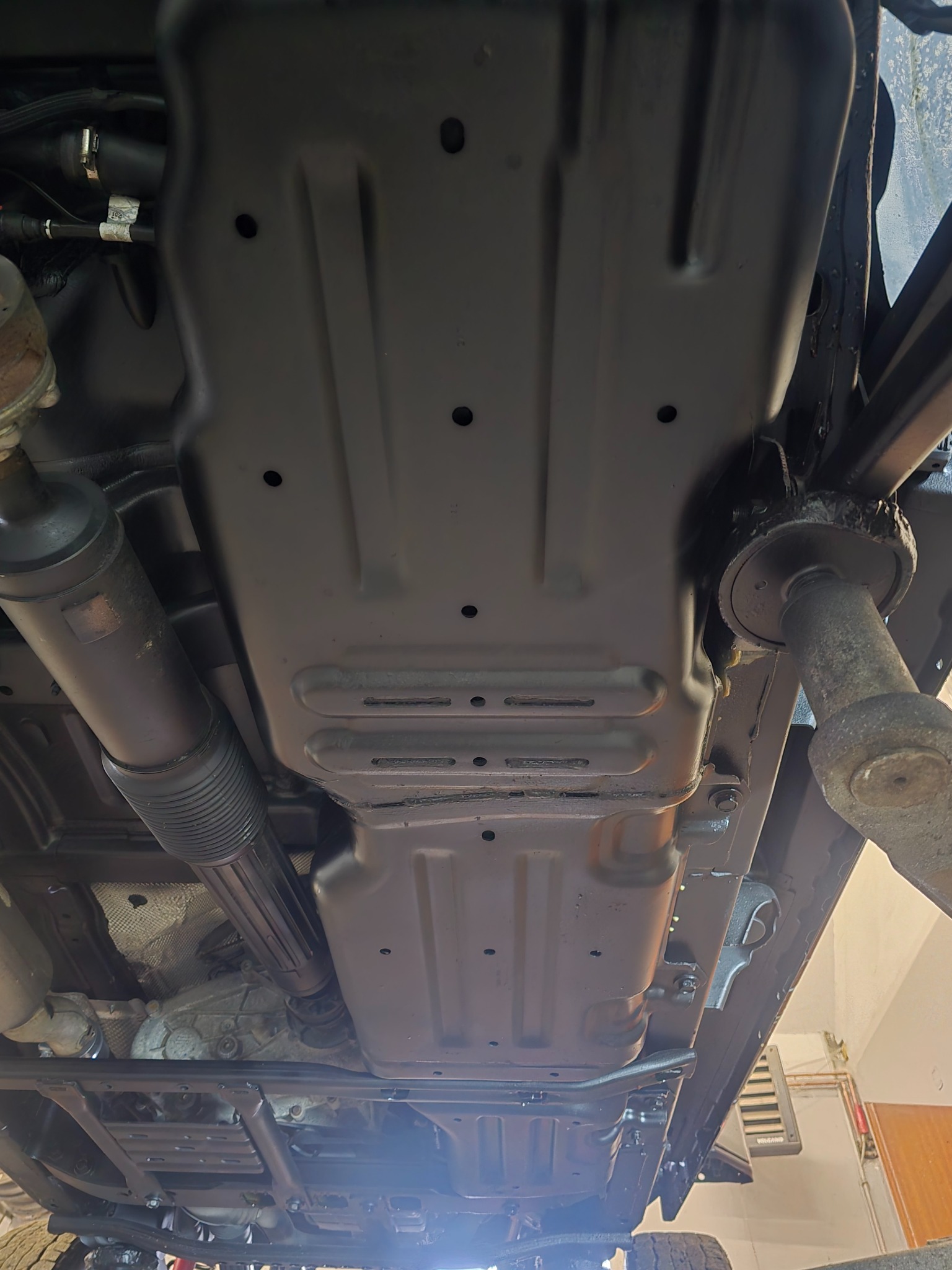 Podwozie samochodu po konserwacji antykorozyjnej, widok od spodu. Elementy metalowe pokryte czarną, ochronną powłoką. Widoczne elementy zawieszenia i układu wydechowego.