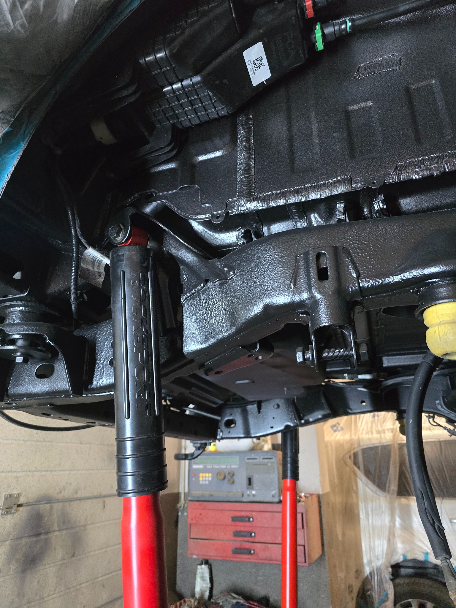 Podwozie auta po konserwacji antykorozyjnej, widoczne elementy zawieszenia, amortyzator RockSport, podnośnik warsztatowy i wyposażenie warsztatu w tle.