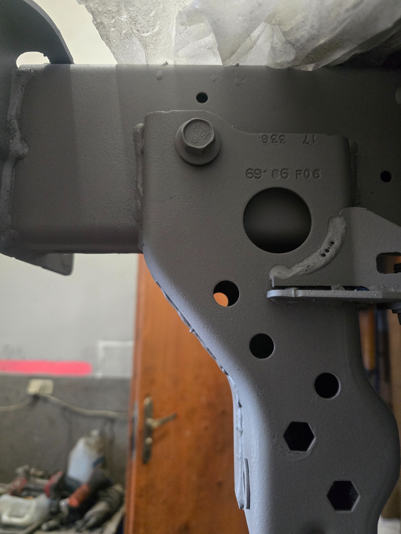 Szczegółowe ujęcie ramy pojazdu po konserwacji antykorozyjnej, z widocznymi oznaczeniami i śrubą. Szary, matowy kolor powłoki. Tło warsztatowe.