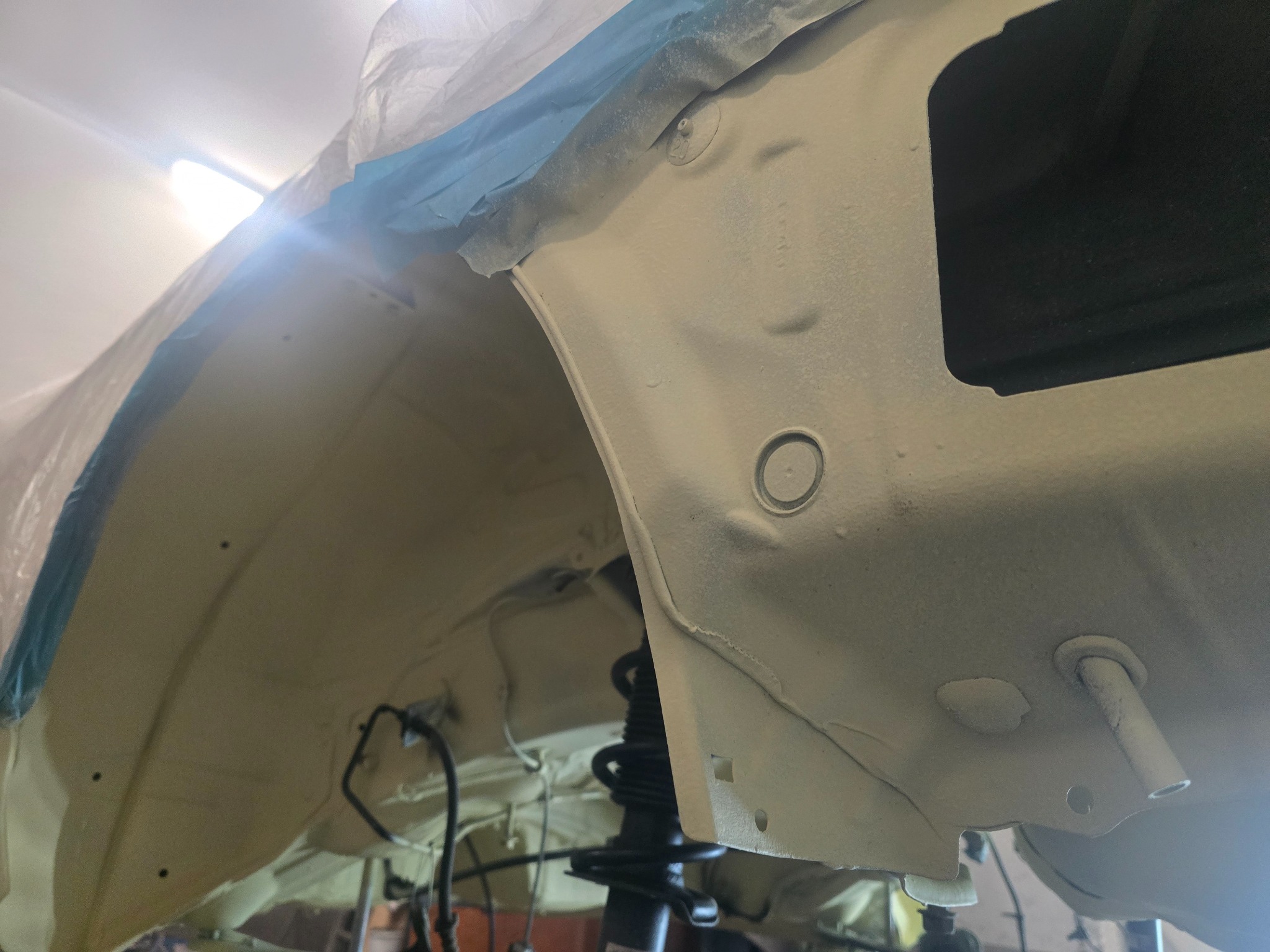 Błotnik pojazdu po konserwacji antykorozyjnej, zabezpieczony taśmą malarską. Widoczna sprężyna zawieszenia i elementy podwozia pokryte jasną farbą ochronną.