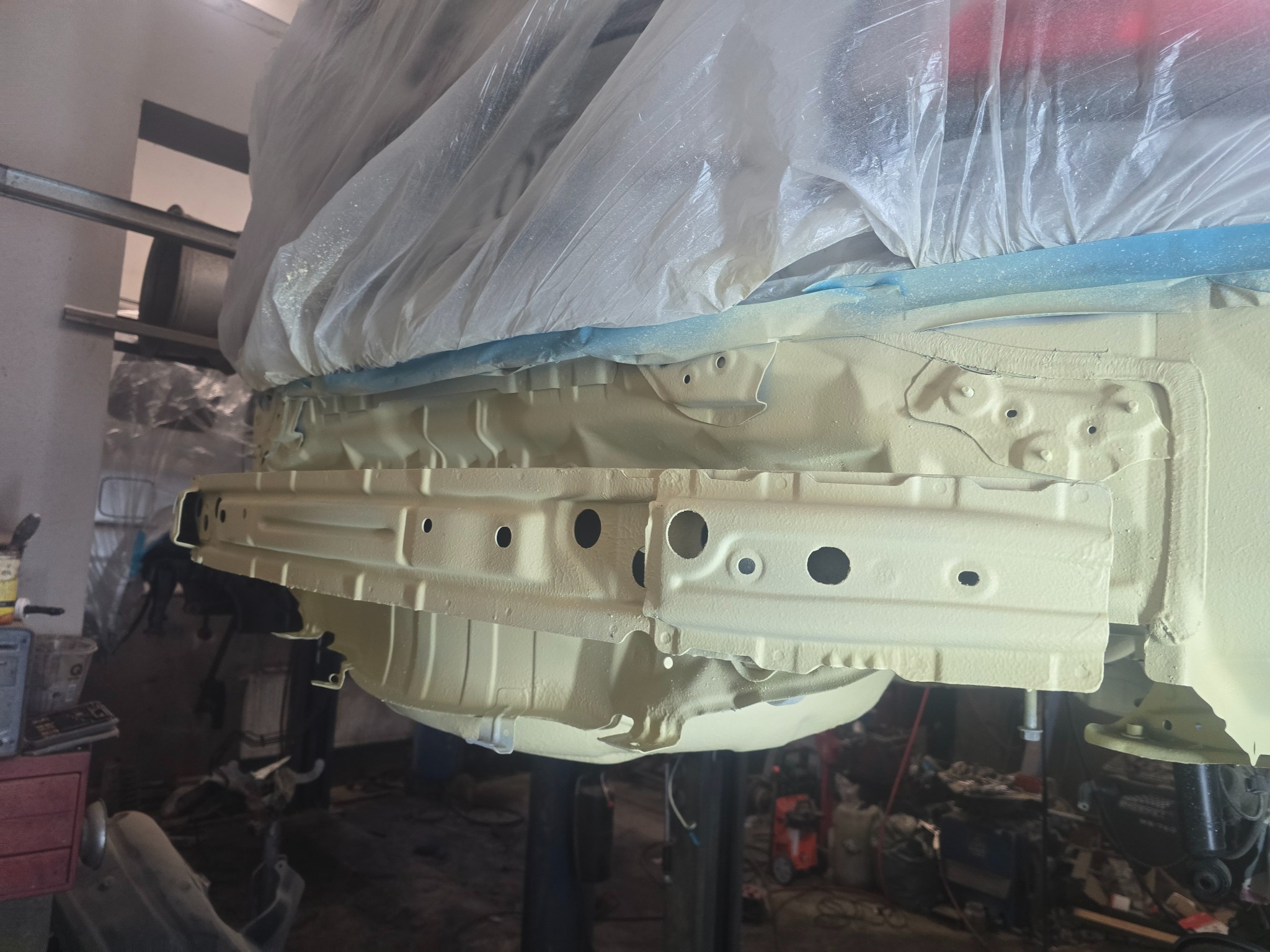 Podwozie samochodu na podnośniku po nałożeniu warstwy ochronnej, zabezpieczone folią i taśmą malarską. Widoczne elementy konstrukcyjne i otwory technologiczne.