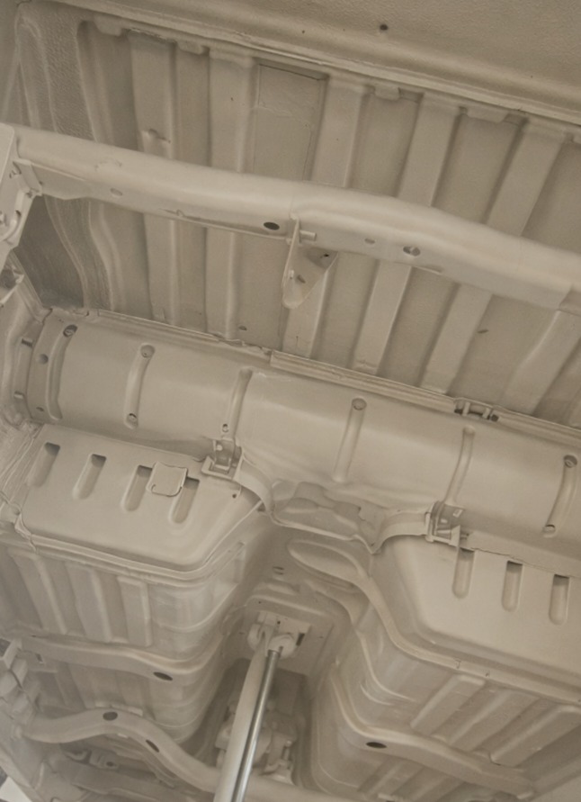 Białe, świeżo pomalowane podwozie samochodu na podnośniku. Widoczne elementy konstrukcyjne i zabezpieczenia antykorozyjne. Jasne oświetlenie warsztatowe.