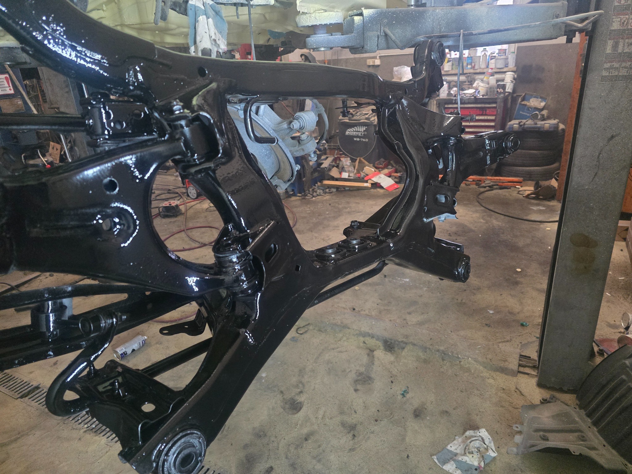 Błyszcząca, czarna rama podwozia samochodu po konserwacji antykorozyjnej, widoczna w warsztacie samochodowym z narzędziami i oponami w tle.