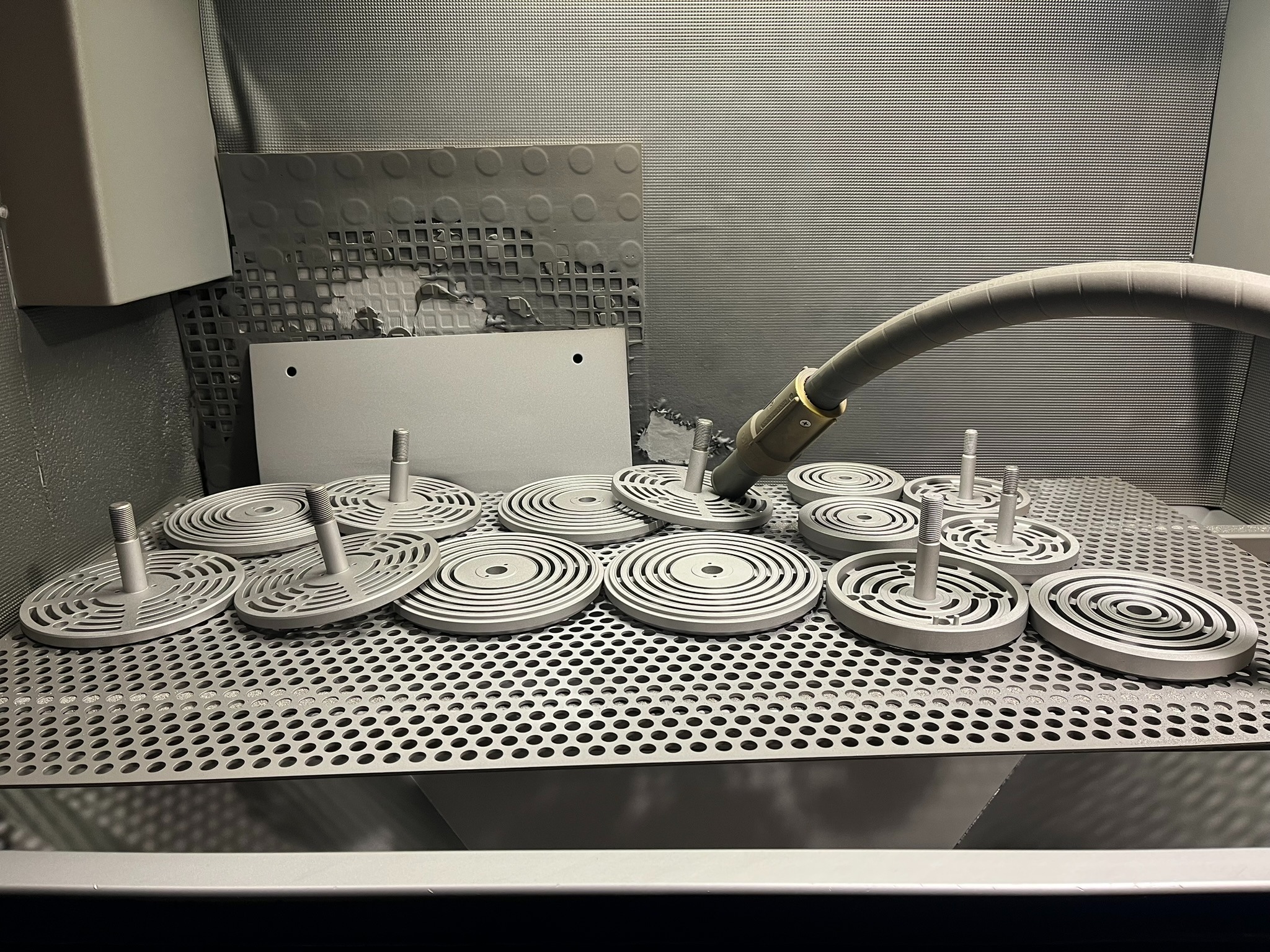 Wnętrze kabiny do obróbki strumieniowo-ściernej z dyszą i detalami w kształcie krążków z centralnym trzpieniem, leżącymi na perforowanym stole.