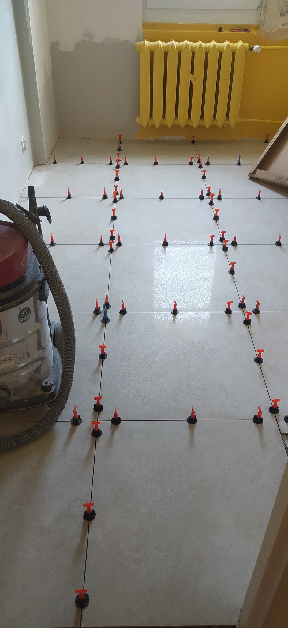 Układanie jasnych płytek podłogowych z użyciem systemu poziomowania. W tle żółty grzejnik i odkurzacz przemysłowy.