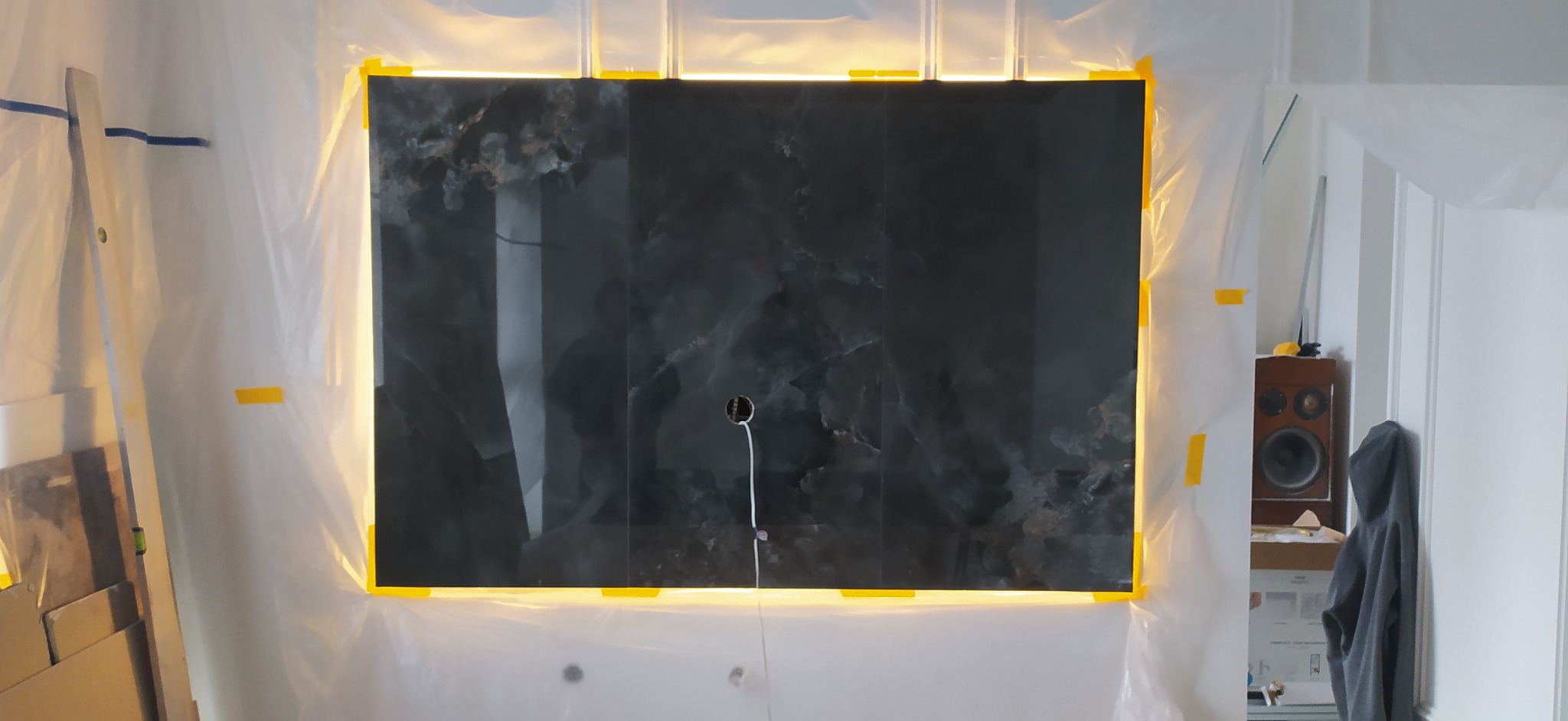 Ściana wyłożona płytami z efektem marmuru, podświetlona taśmą LED. Zabezpieczona folią malarską. Widoczny głośnik w tle.