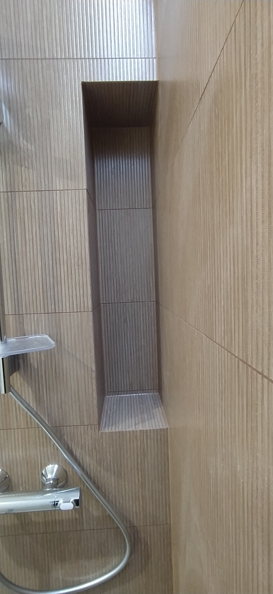 Beżowa ściana z płytek imitujących drewno w łazience, z wnęką wyłożoną szarymi płytkami o pionowym wzorze. Widoczny fragment baterii prysznicowej.