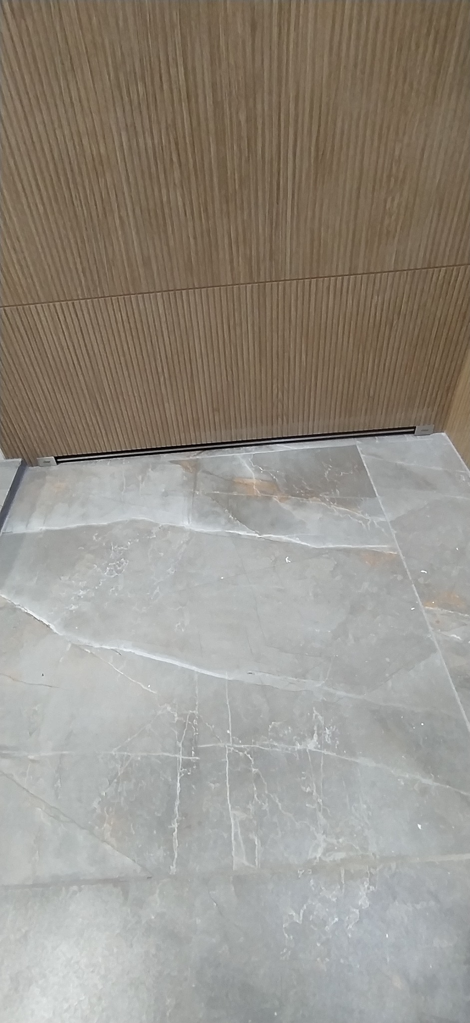 Stylowe wykończenie: szare płytki podłogowe imitujące marmur i panel ścienny z subtelnym, prążkowanym wzorem drewna. Minimalistyczna listwa przypodłogowa.