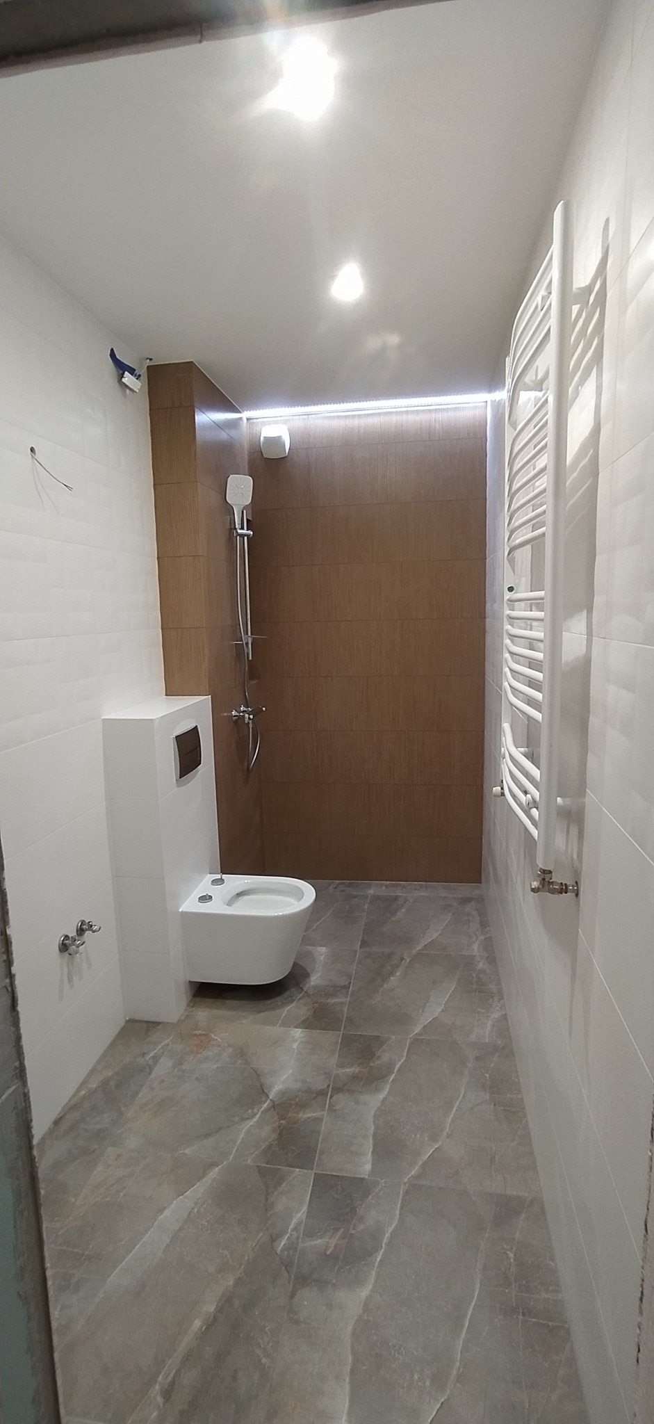 Nowoczesna łazienka po remoncie: podwieszana toaleta, brązowe płytki prysznicowe z oświetleniem LED i marmurowe płytki podłogowe. Grzejnik drabinkowy.