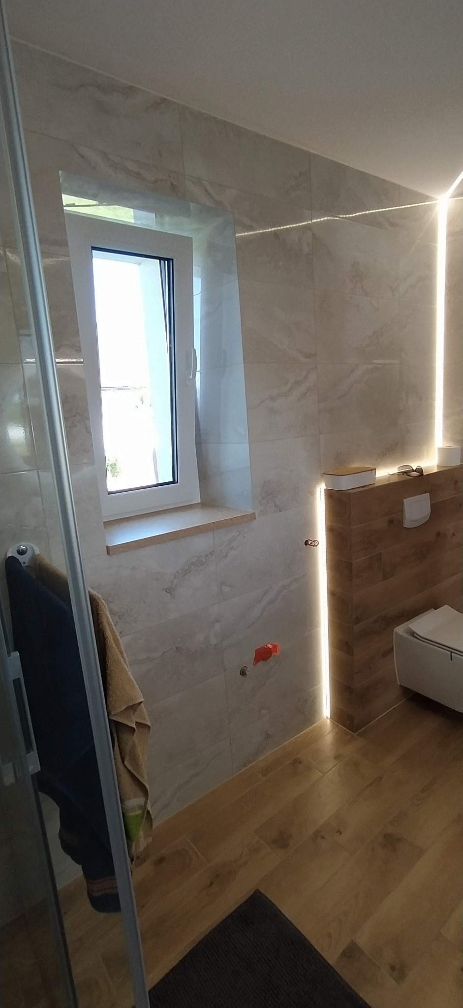 Nowoczesna łazienka z oknem, beżowymi płytkami imitującymi marmur i drewnianymi akcentami. Uwagę przykuwa podświetlenie LED oraz wisząca miska WC.