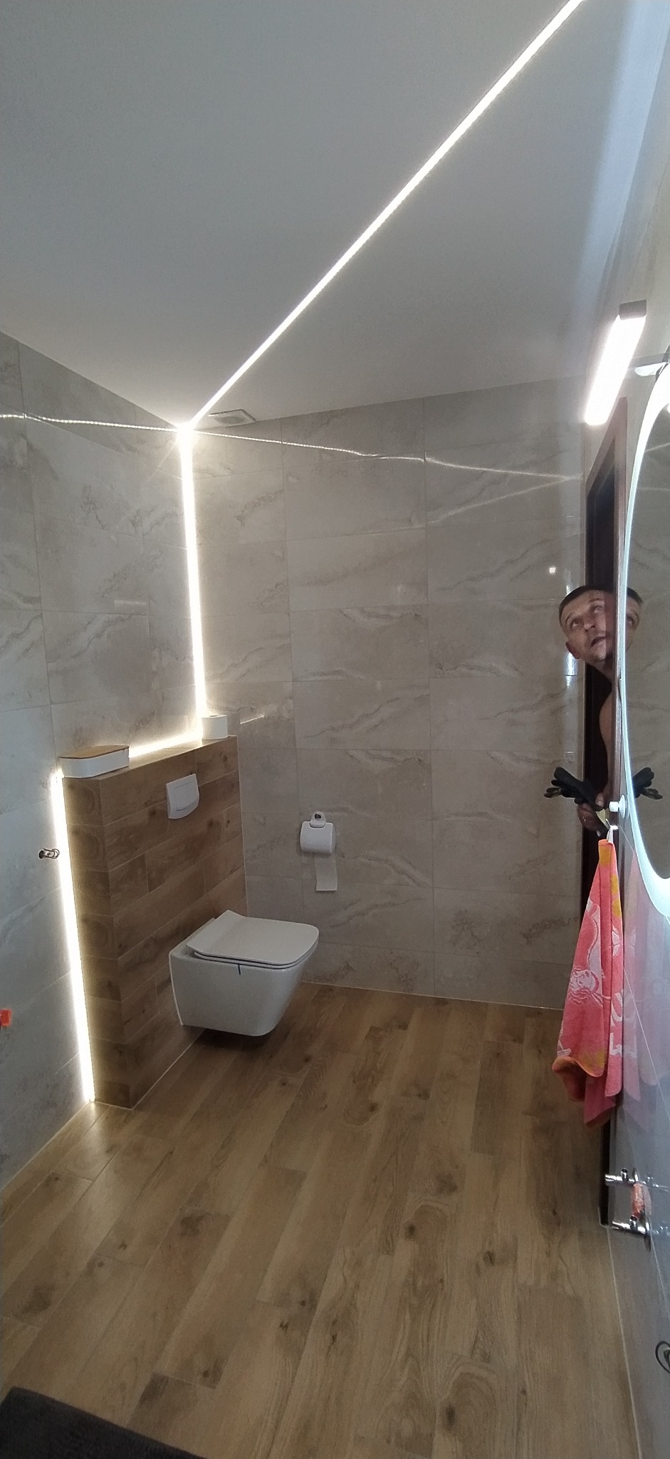 Nowoczesna łazienka z podwieszaną toaletą, drewnopodobną podłogą i marmurowymi płytkami. Oświetlenie LED podkreśla aranżację. Osoba w odbiciu lustra.