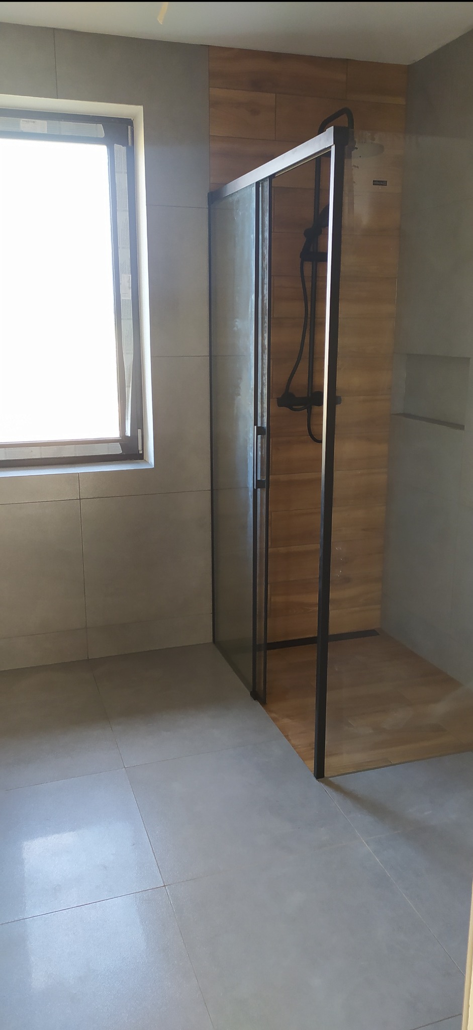 Nowoczesna kabina prysznicowa z czarnymi profilami i drewnopodobnymi płytkami wewnątrz, na tle szarych płytek ściennych i podłogowych. Okno z widokiem na zewnątrz.