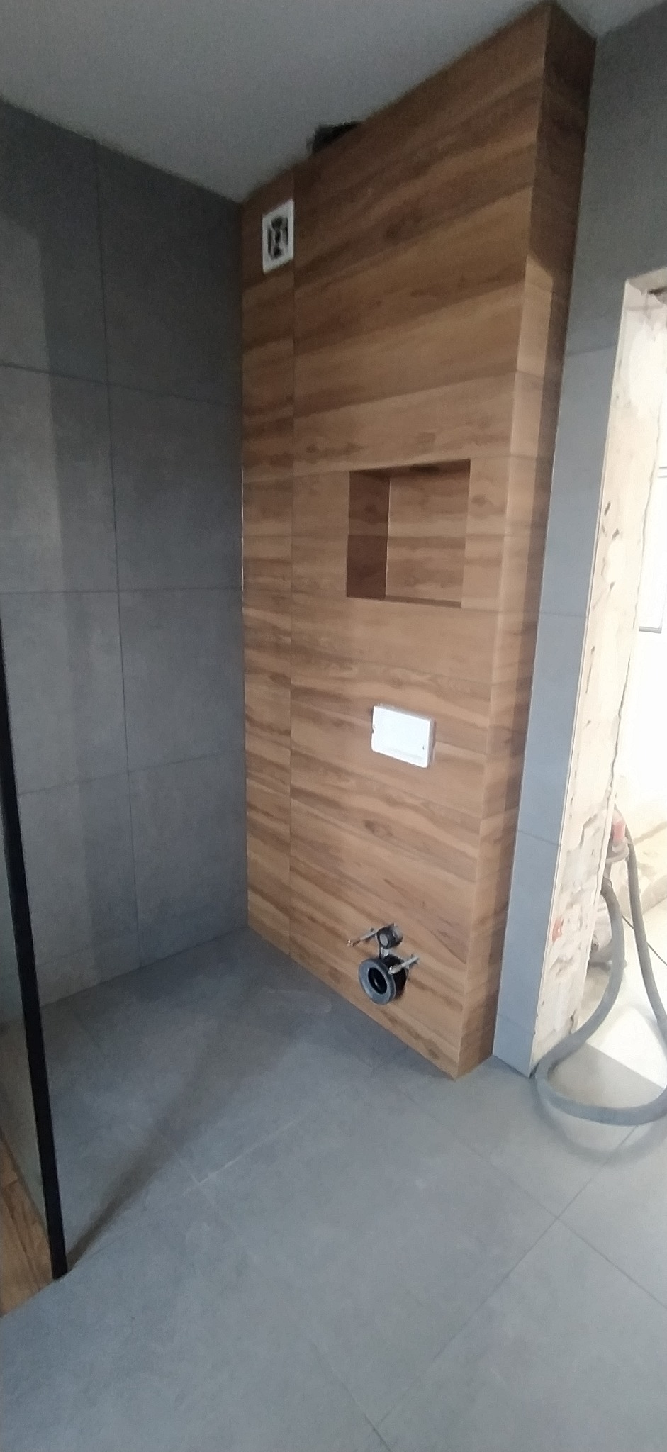 Nowoczesna łazienka w trakcie wykańczania: drewnopodobne płytki na ścianie z wnęką, szare płytki podłogowe i ścienne. Widoczne elementy instalacji podtynkowej WC.