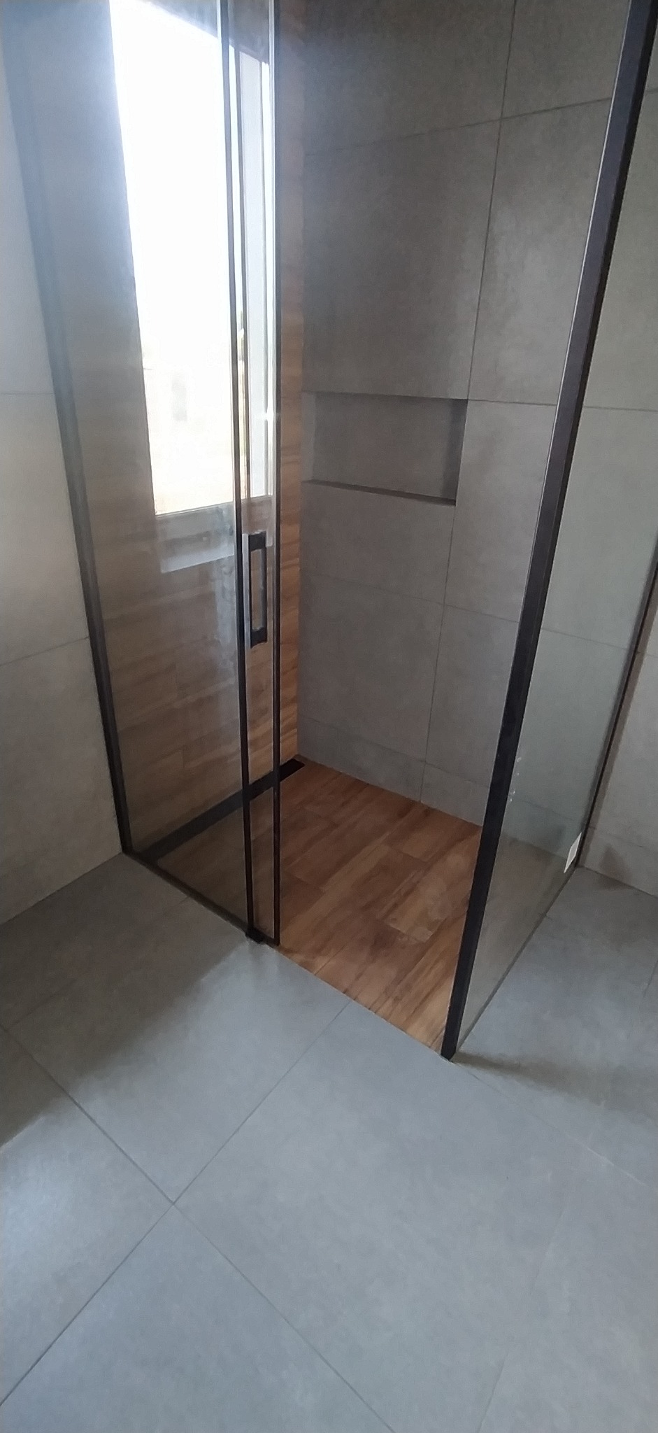 Nowoczesna kabina prysznicowa z drewnopodobną podłogą i szarymi płytkami. Widoczne detale wykończenia i minimalistyczny design.