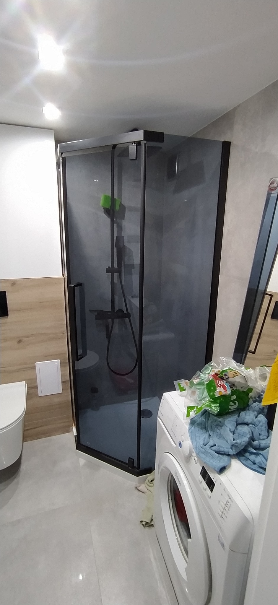 Nowoczesna kabina prysznicowa z czarnymi profilami w wykończonej łazience. Widoczna toaleta i pralka. Minimalistyczny design, szare płytki podłogowe.