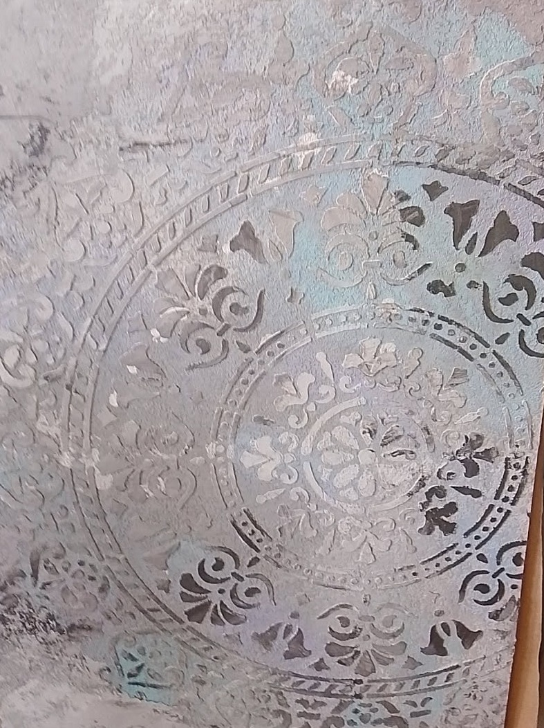 Dekoracyjna ściana w odcieniach szarości z motywem mandali, wykonana techniką szablonową. Widoczne niedoskonałości faktury i subtelne przebarwienia błękitu.