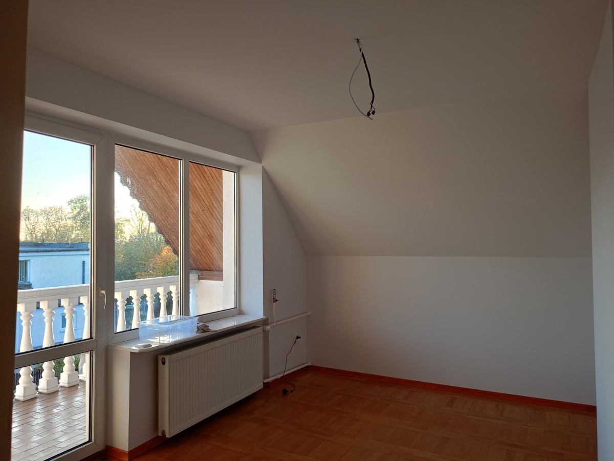 Pokój na poddaszu z oknem balkonowym i parkietem. Ściany pomalowane na biało, widoczne przewody elektryczne. Na zewnątrz balkon z białą balustradą.