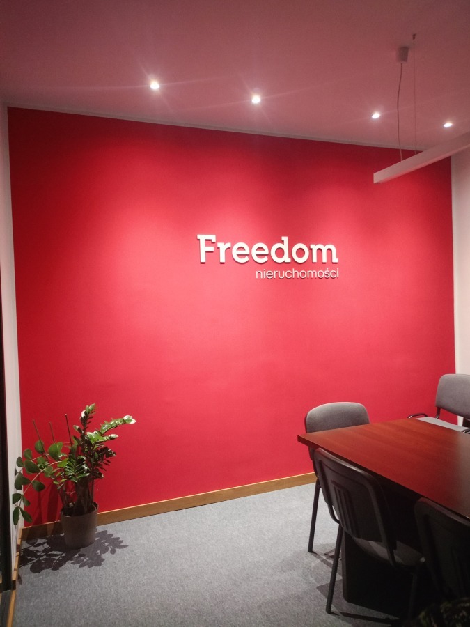 Czerwona ściana z białym napisem 'Freedom nieruchomości' w biurze. Na podłodze szary dywan, w rogu doniczka z rośliną, widoczne krzesła i stół.