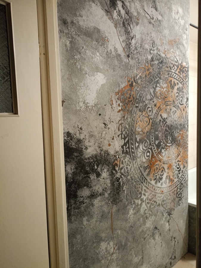 Dekoracyjna ściana z efektem betonu i ornamentem w łazience, widoczna obok białych drzwi i fragmentu wanny. Nietypowe wykończenie z bliska.