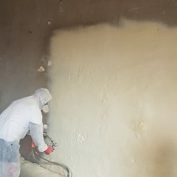 PURIKA Dawid Koszyka - Pracownik w kombinezonie ochronnym natryskuje pianę izolacyjną na ścianę, widoczny fragment ściany przed i po ociepleniu.