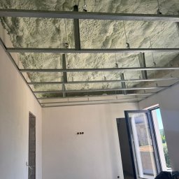 PURIKA Dawid Koszyka - Poddasze po ociepleniu pianką PUR, widoczna metalowa konstrukcja sufitu podwieszanego i nałożona pianka. Częściowo otwarte okno i worki ze śmieciami na podłodze.