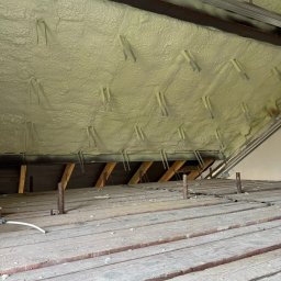 PURIKA Dawid Koszyka - Poddasze ocieplone pianką PUR, widoczne drewniane belki i metalowe elementy mocujące, jasna piana szczelnie wypełnia przestrzeń między krokwiami.