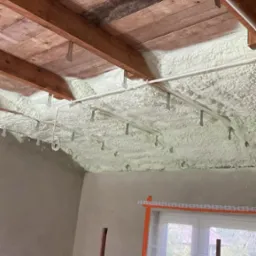 Poddasze z drewnianą konstrukcją i ociepleniem pianką PUR, widoczne elementy instalacji i zabezpieczone okno folią. Fragment rusztowania w kadrze.