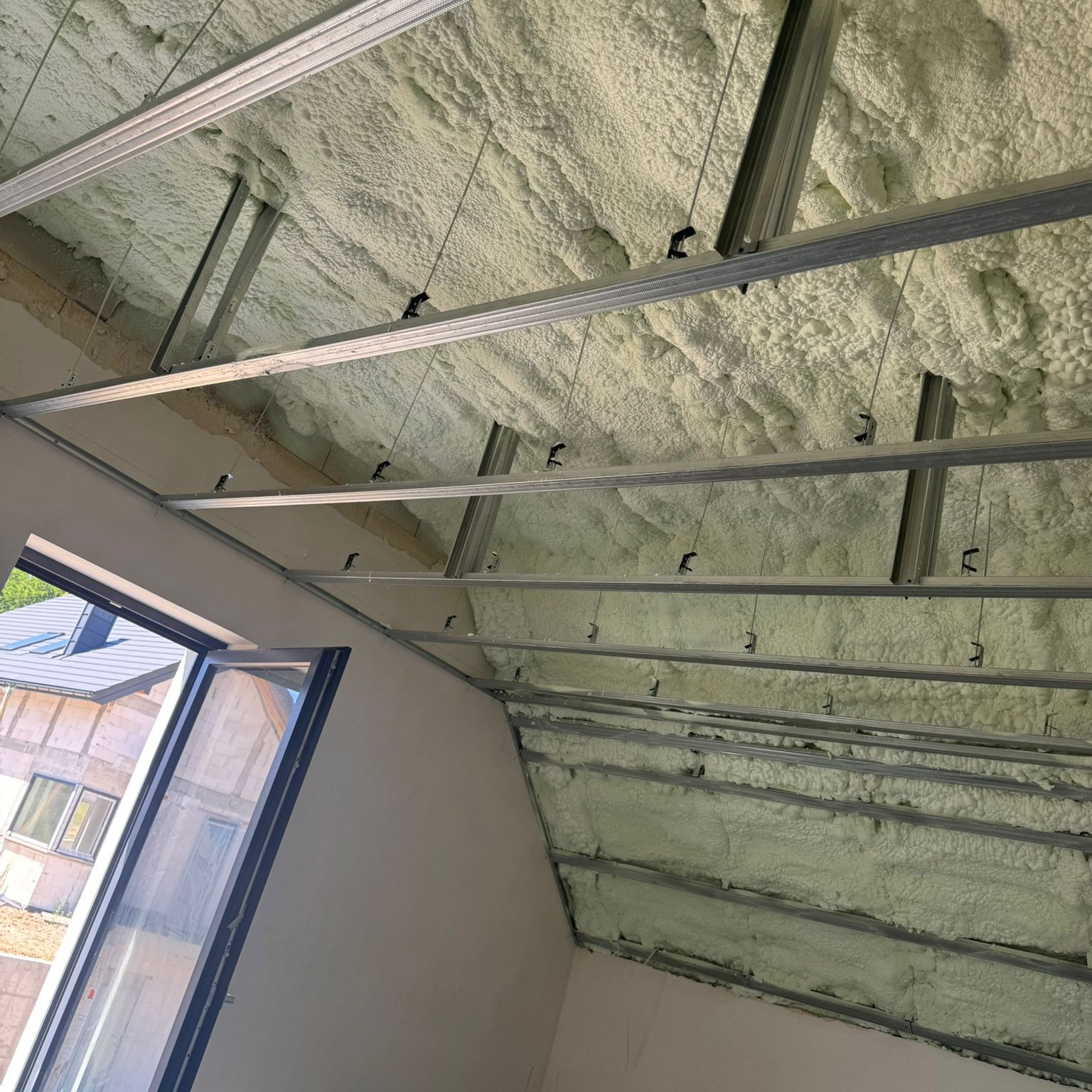 Poddasze z ociepleniem pianką PUR, widoczne metalowe profile konstrukcyjne. Ujęcie z dołu, w tle okno dachowe i fragment budynku.