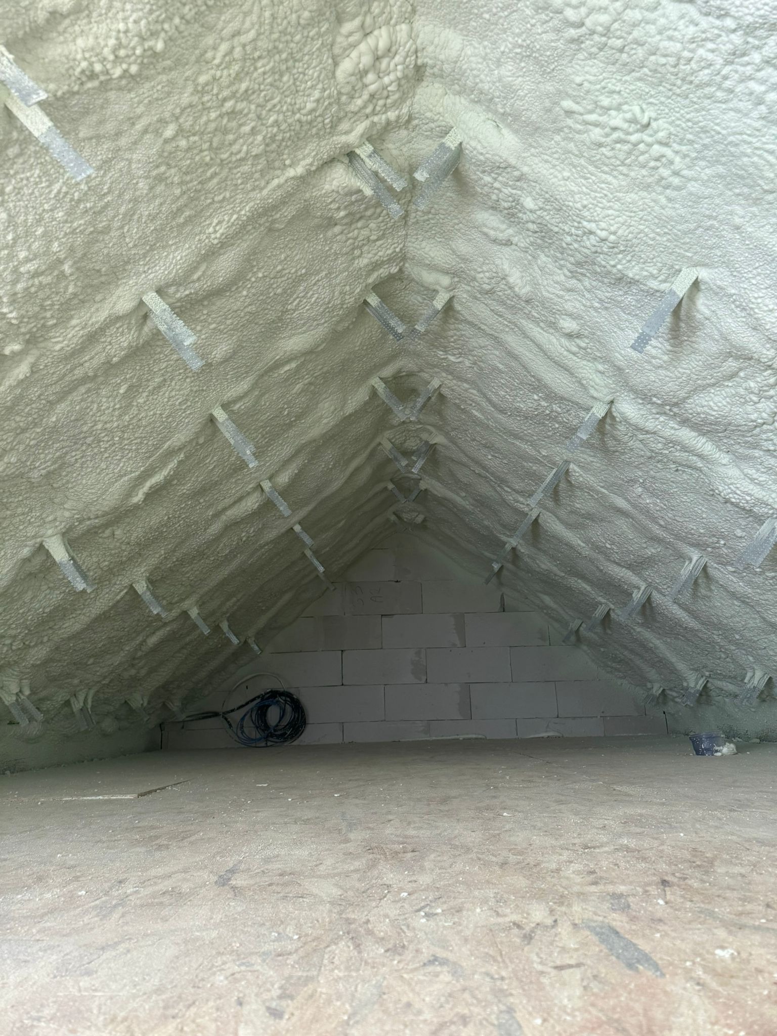 Poddasze ocieplone pianką PUR, widoczne krokwie i biała pianka, z przewodami elektrycznymi przy ścianie z bloczków. Wykończona podłoga.