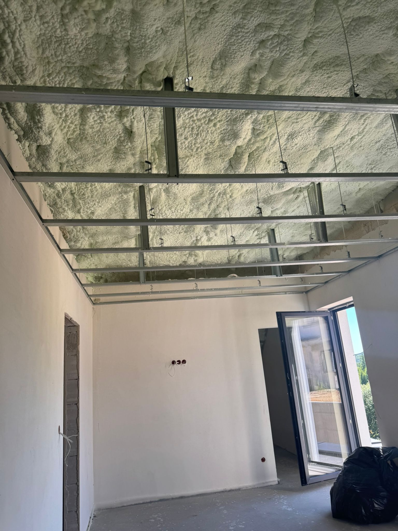 Poddasze po ociepleniu pianką PUR, widoczna metalowa konstrukcja sufitu podwieszanego i nałożona pianka. Częściowo otwarte okno i worki ze śmieciami na podłodze.