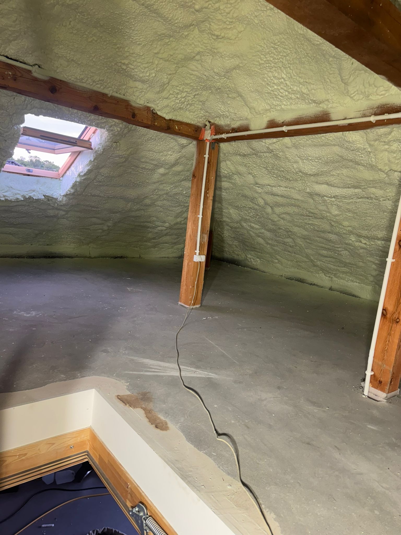 Poddasze po ociepleniu pianką PUR, widoczne drewniane elementy konstrukcyjne i okno dachowe. Szara podłoga, białe rury, oświetlenie dzienne.