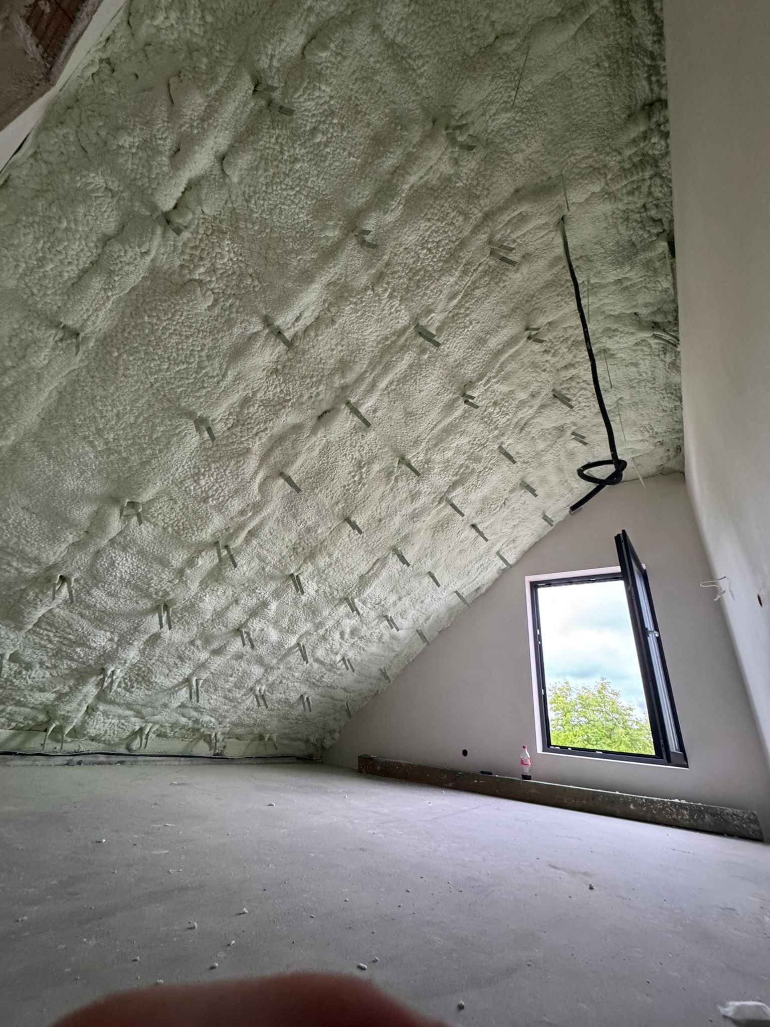 Poddasze po ociepleniu pianką PUR, widoczne szpilki mocujące izolację, okno dachowe z widokiem na zieleń. Kabel elektryczny zwisa z sufitu.