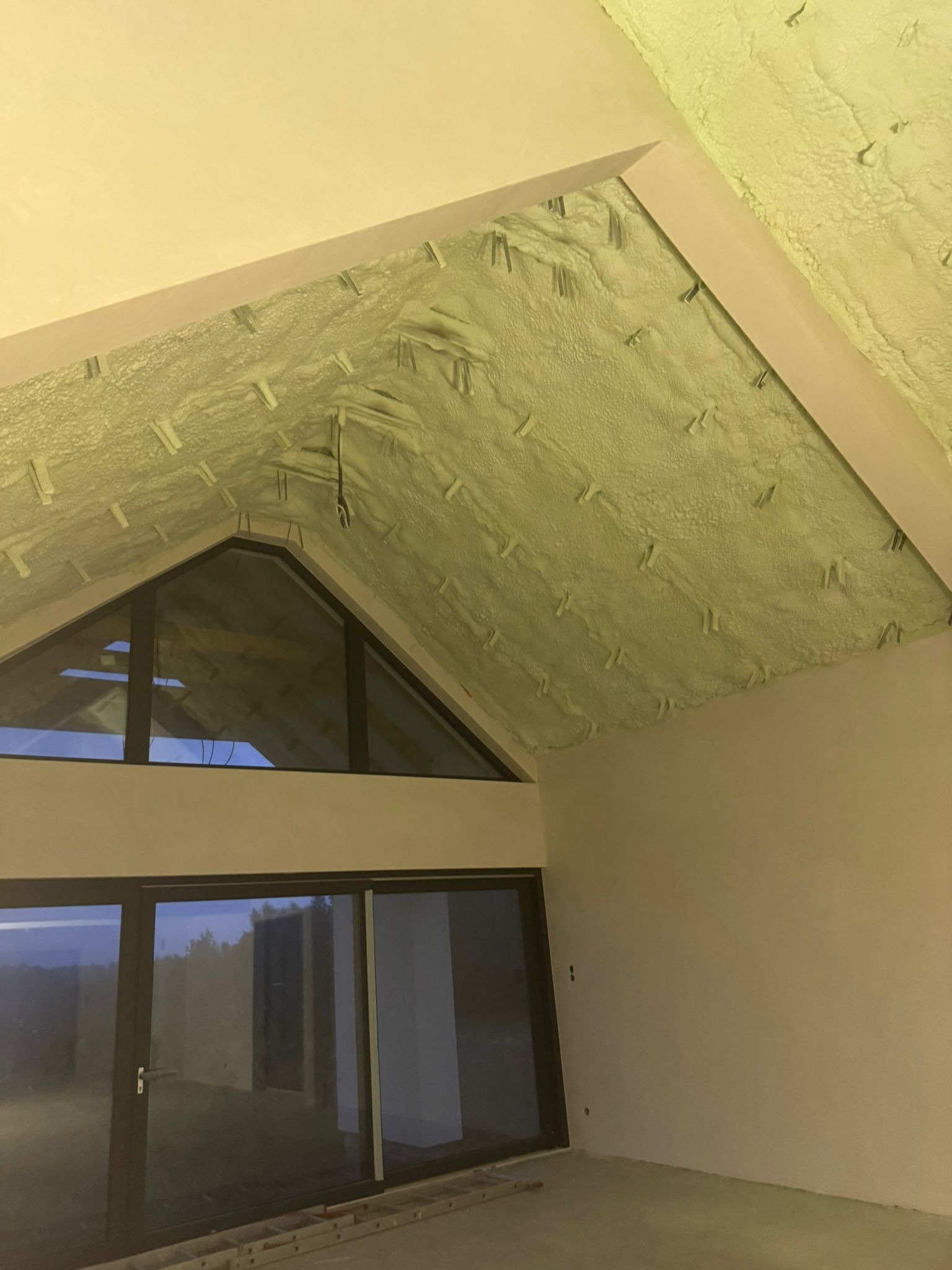 Poddasze po ociepleniu pianką PUR, widoczne mocowania i fragment okna. Ściany w jasnym odcieniu, na podłodze drabina.