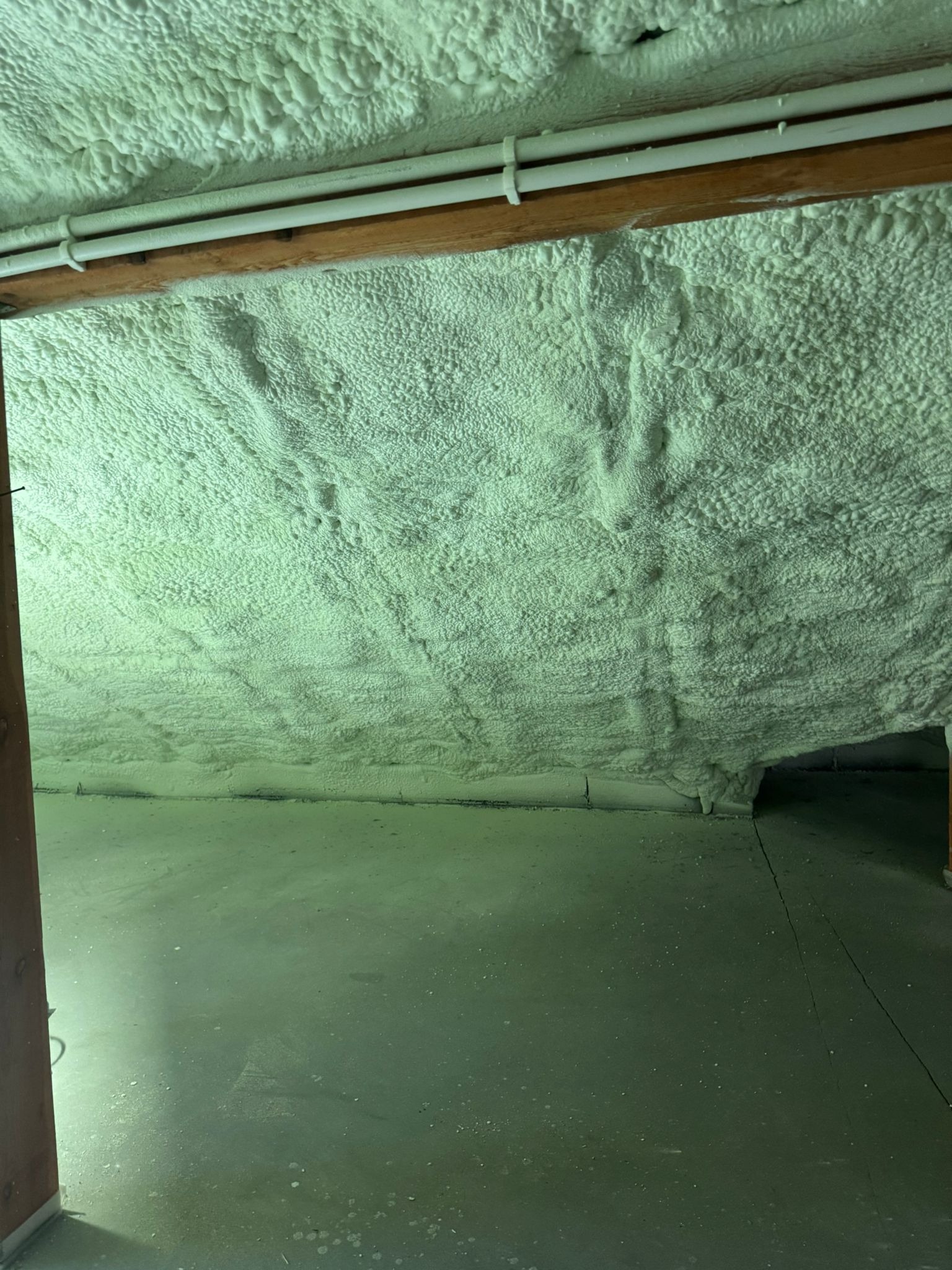 Poddasze po ociepleniu pianką PUR; widoczne białe rury i drewniana belka. Ściany i sufit pokryte jednolitą warstwą pianki, szara podłoga.