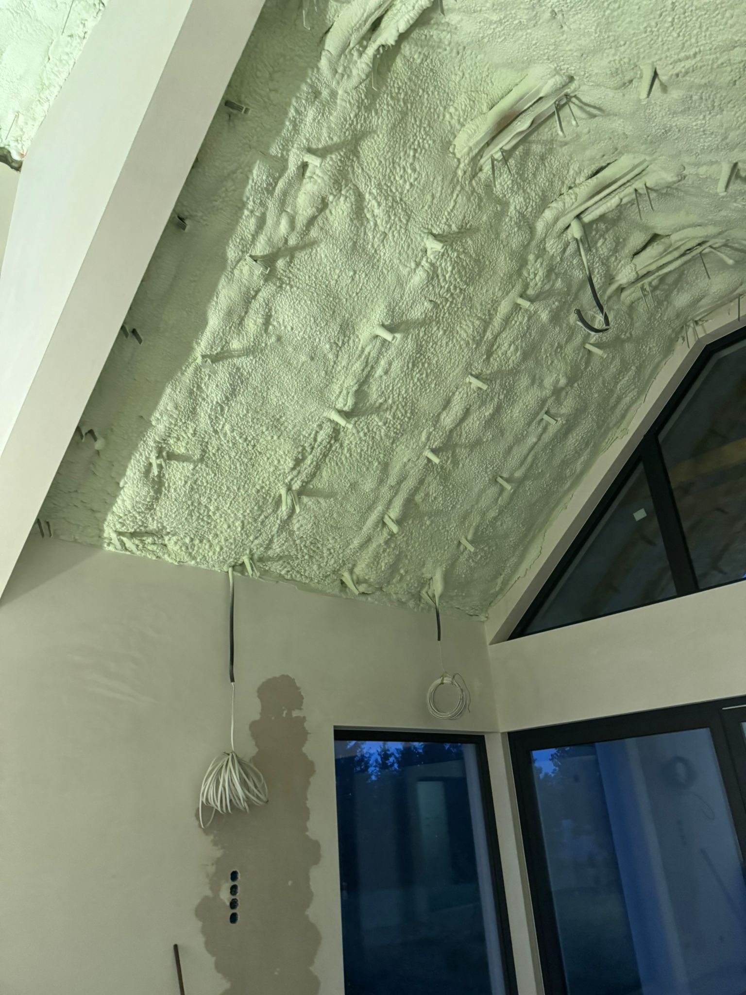 Poddasze po ociepleniu pianką PUR, widoczne wystające elementy montażowe. Ściany w trakcie prac wykończeniowych, okna z ciemnymi ramami, przewody elektryczne.