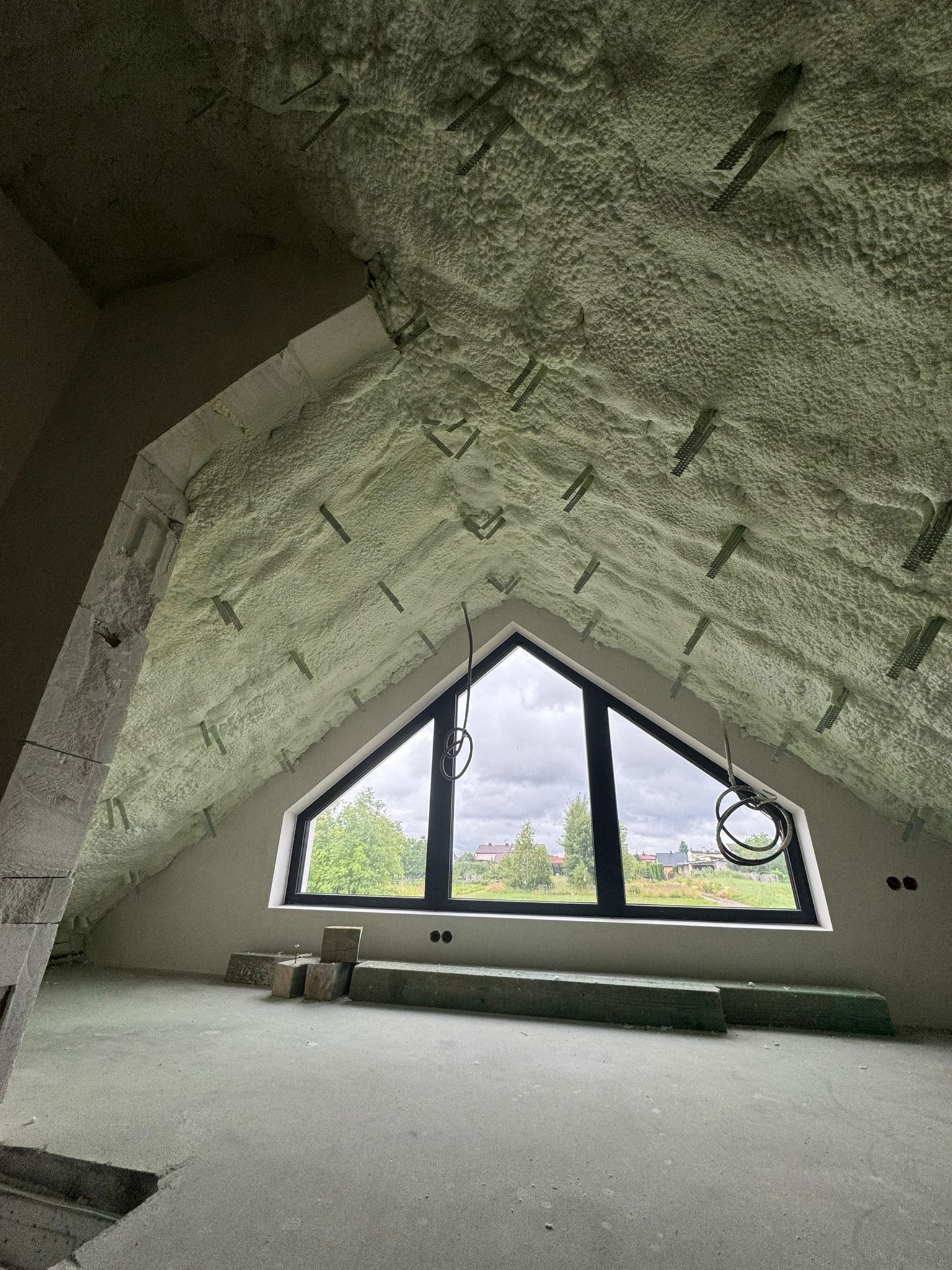 Poddasze po ociepleniu pianką PUR, widoczne elementy montażowe. Okno trójkątne z widokiem na zewnątrz. Surowy stan wykończenia podłogi i ścian.