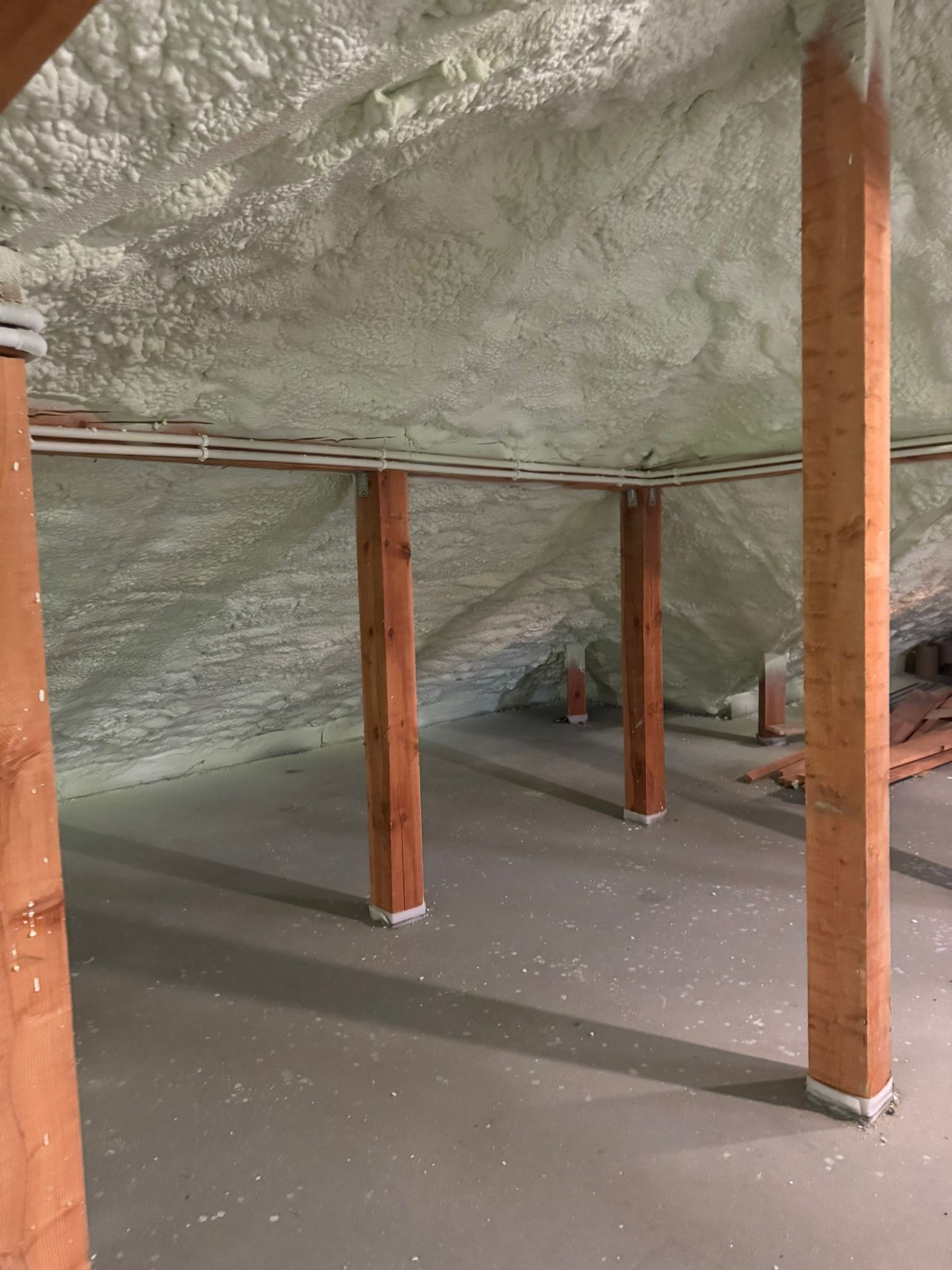 Poddasze po ociepleniu pianką PUR, widoczne drewniane słupy i instalacje. Biała, nierówna powierzchnia pianki pokrywa skośny sufit i ściany.