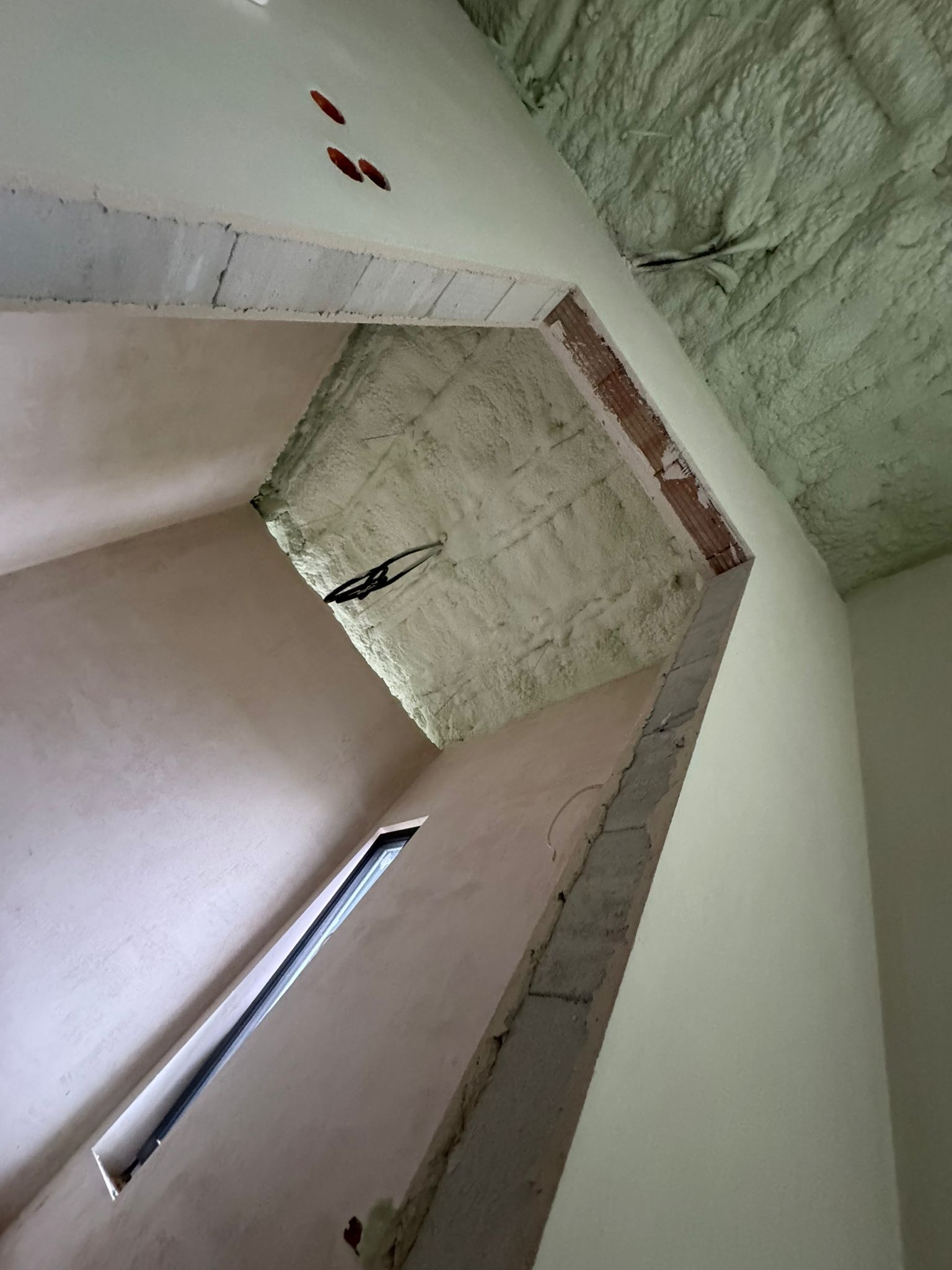 Poddasze po ociepleniu pianką PUR, widoczne nierówności piany na suficie, okno w ścianie, surowe wykończenie ścian, kabel zwisający z sufitu.