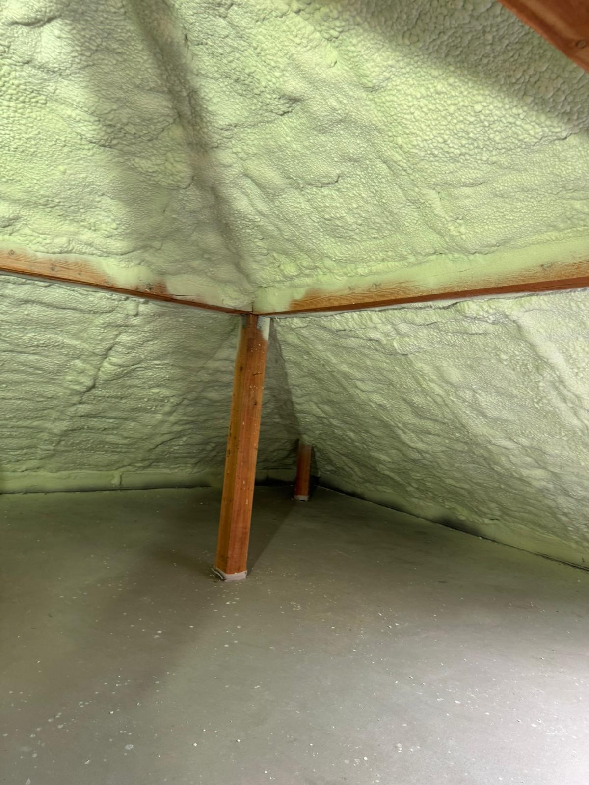 Poddasze po ociepleniu pianką PUR, widoczne drewniane elementy konstrukcyjne i szara podłoga. Pianka ma jasny, zielonkawy odcień i nierównomierną strukturę.