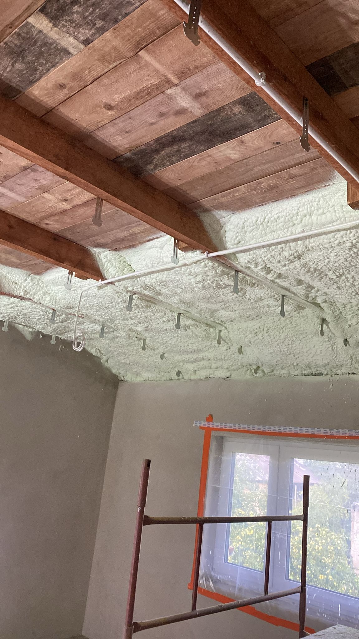 Poddasze z drewnianą konstrukcją i ociepleniem pianką PUR, widoczne elementy instalacji i zabezpieczone okno folią. Fragment rusztowania w kadrze.