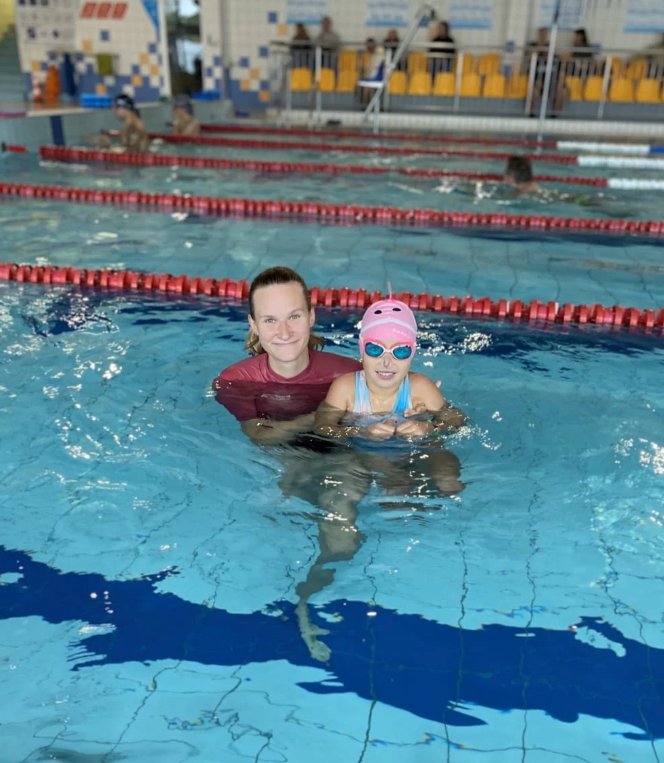 Instruktorka z uśmiechem i dziewczynka w różowym czepku i okularach pływackich w basenie w Poznaniu, podczas lekcji pływania.