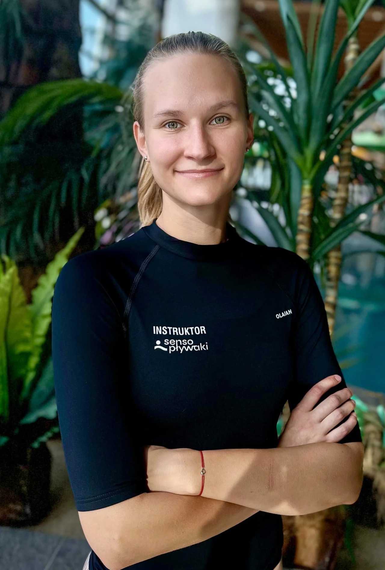 Portret uśmiechniętej instruktorki w czarnym stroju z logo 'Instruktor senso pływaki', na tle tropikalnych roślin w przestrzeni basenowej.