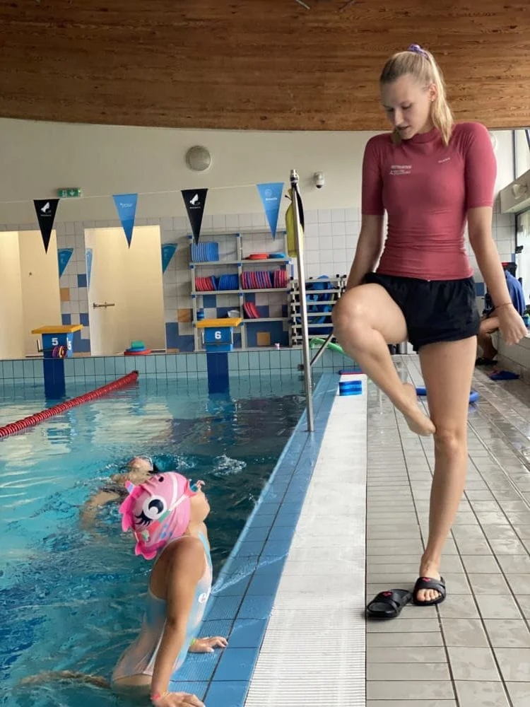 Instruktorka w stroju pływackim demonstruje ćwiczenie rozciągające nogę na brzegu basenu, podczas gdy dziewczynka w czepku z jednorożcem obserwuje ją z wody.