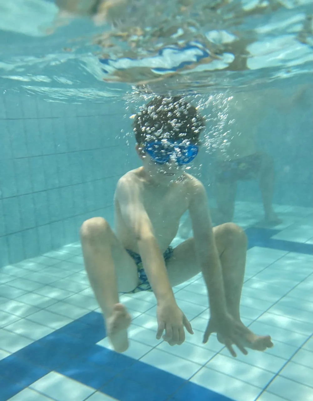 Chłopiec w okularach do pływania zanurzony w basenie, widok z dołu, bąbelki powietrza wokół głowy, płytki basenowe i fragment osoby w tle.