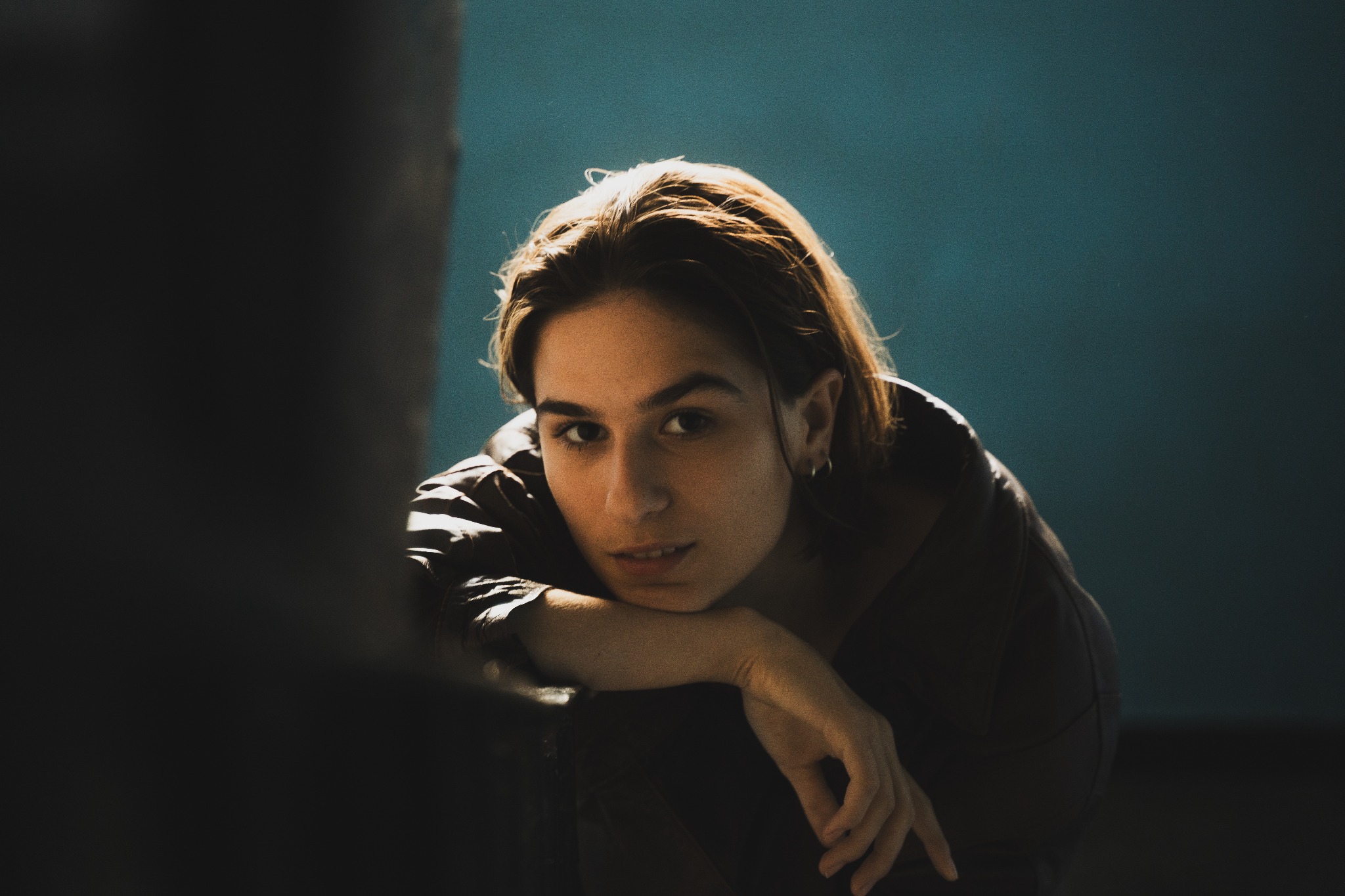 Portret młodej kobiety z krótkimi włosami, opierającej się na ramieniu, z subtelnym uśmiechem i kolczykiem w uchu, na tle niebieskiej ściany z mocnym bocznym oświetleniem.