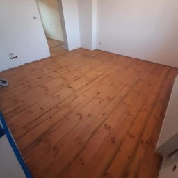 Joanna Staszkiewicz - Wycyklinowana, drewniana podłoga w pokoju z surowymi ścianami, widoczne ślady po cyklinowaniu i fragment stopy w skarpetce.