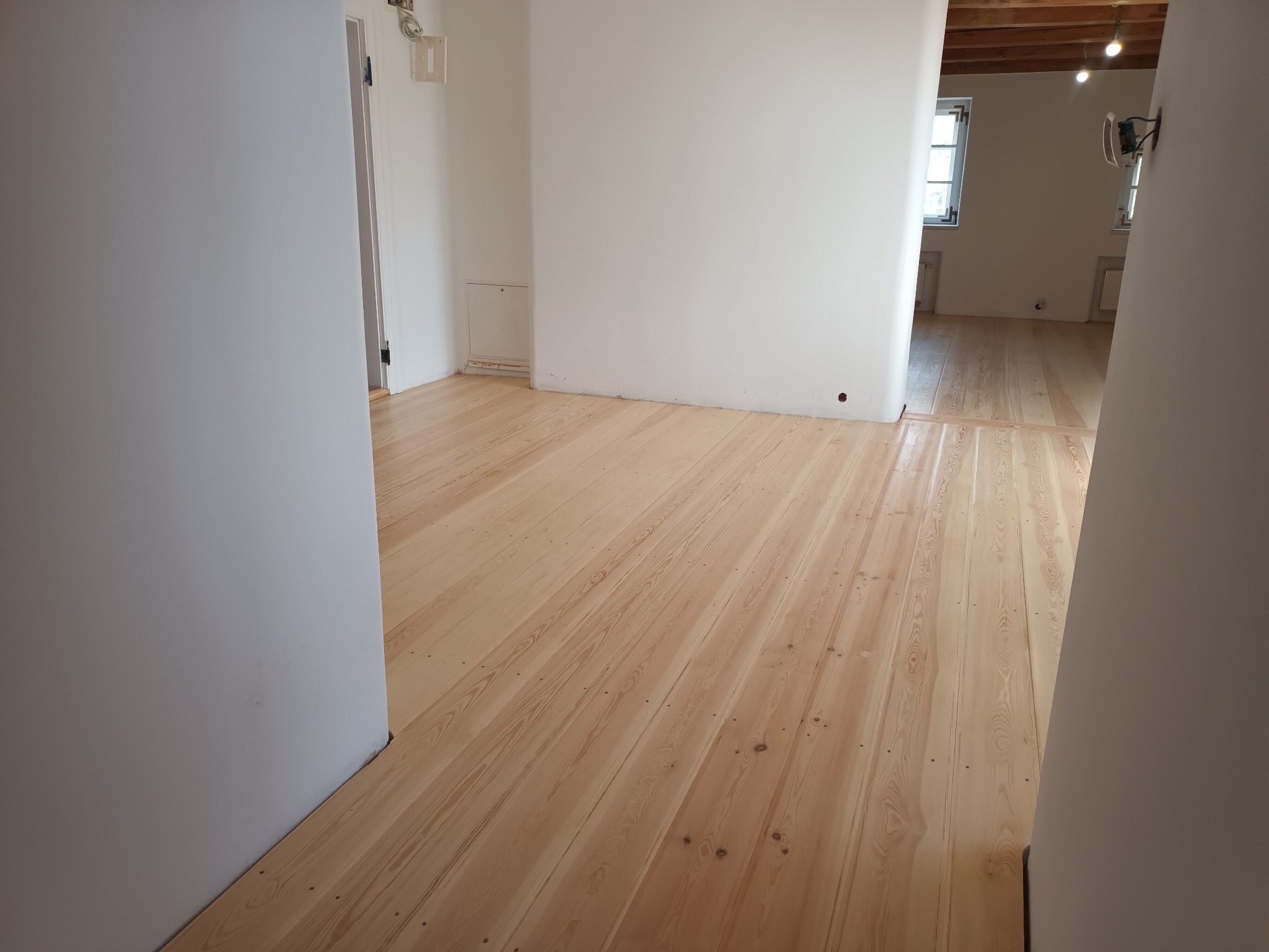 Jasne, drewniane deski podłogowe, świeżo położone, widoczne w pustym, jasnym pokoju z białymi ścianami i drewnianym stropem w tle.