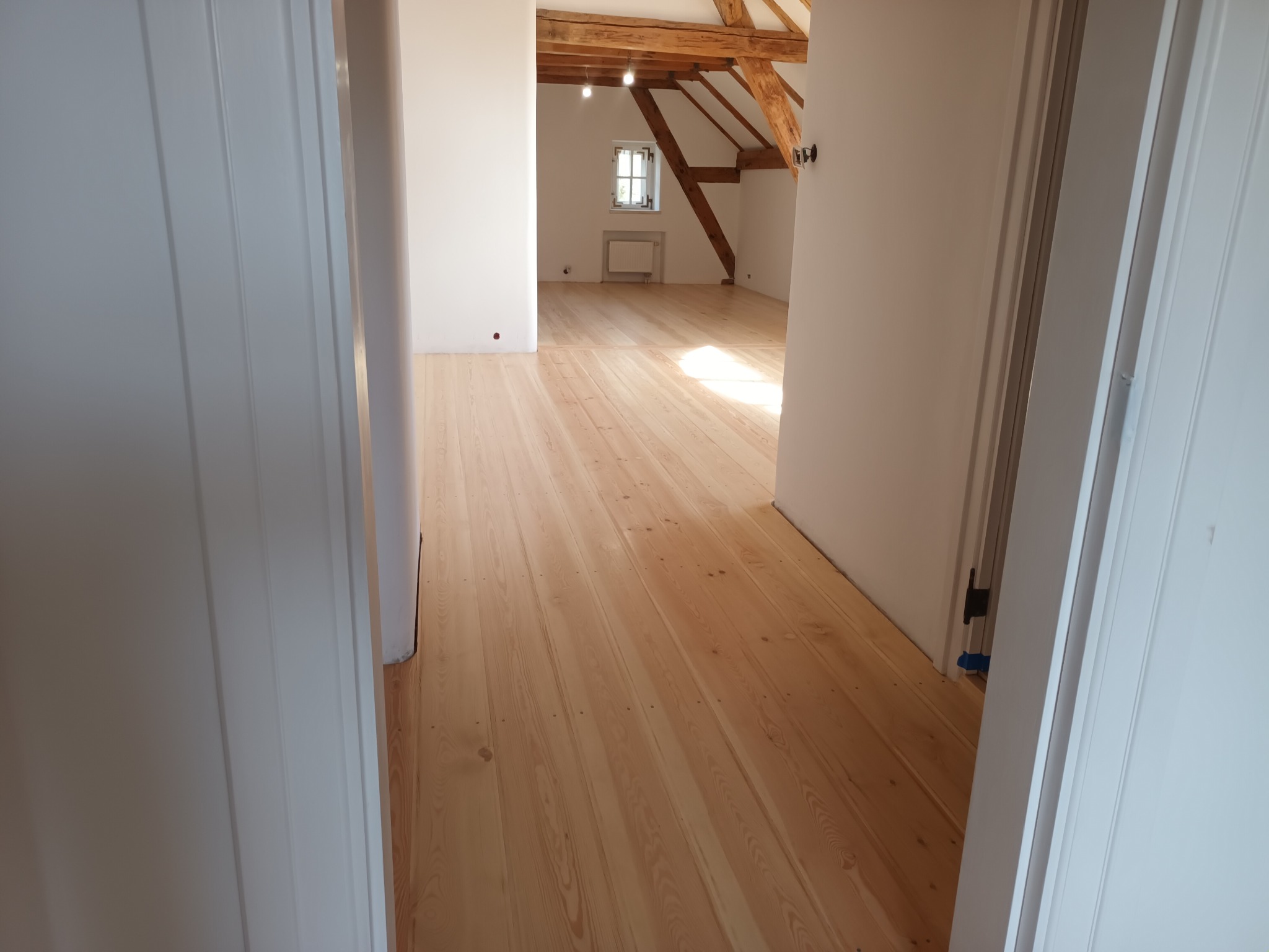 Jasne, drewniane deski podłogowe w poddaszowym pokoju z widoczną więźbą dachową, świeżo po renowacji, widziane zza otwartych, białych drzwi.