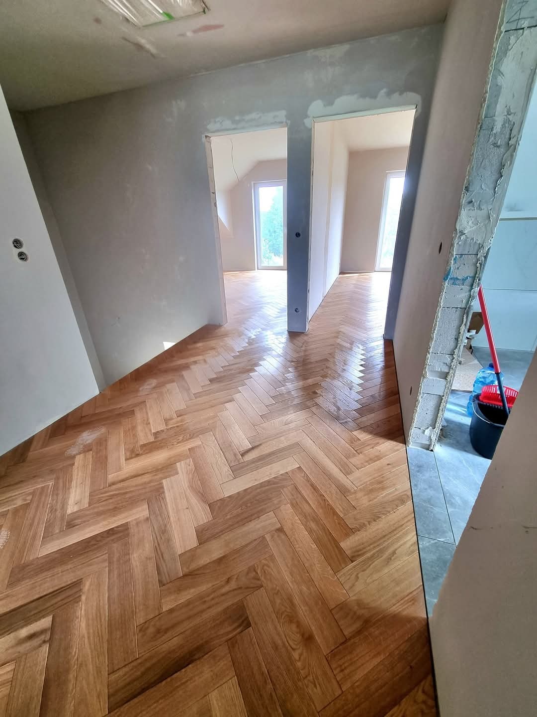 Nowo ułożony parkiet w jodełkę w przestronnym, remontowanym wnętrzu z widocznymi, surowymi ścianami i otworami drzwiowymi prowadzącymi do kolejnych pomieszczeń, gdzie widać okna.