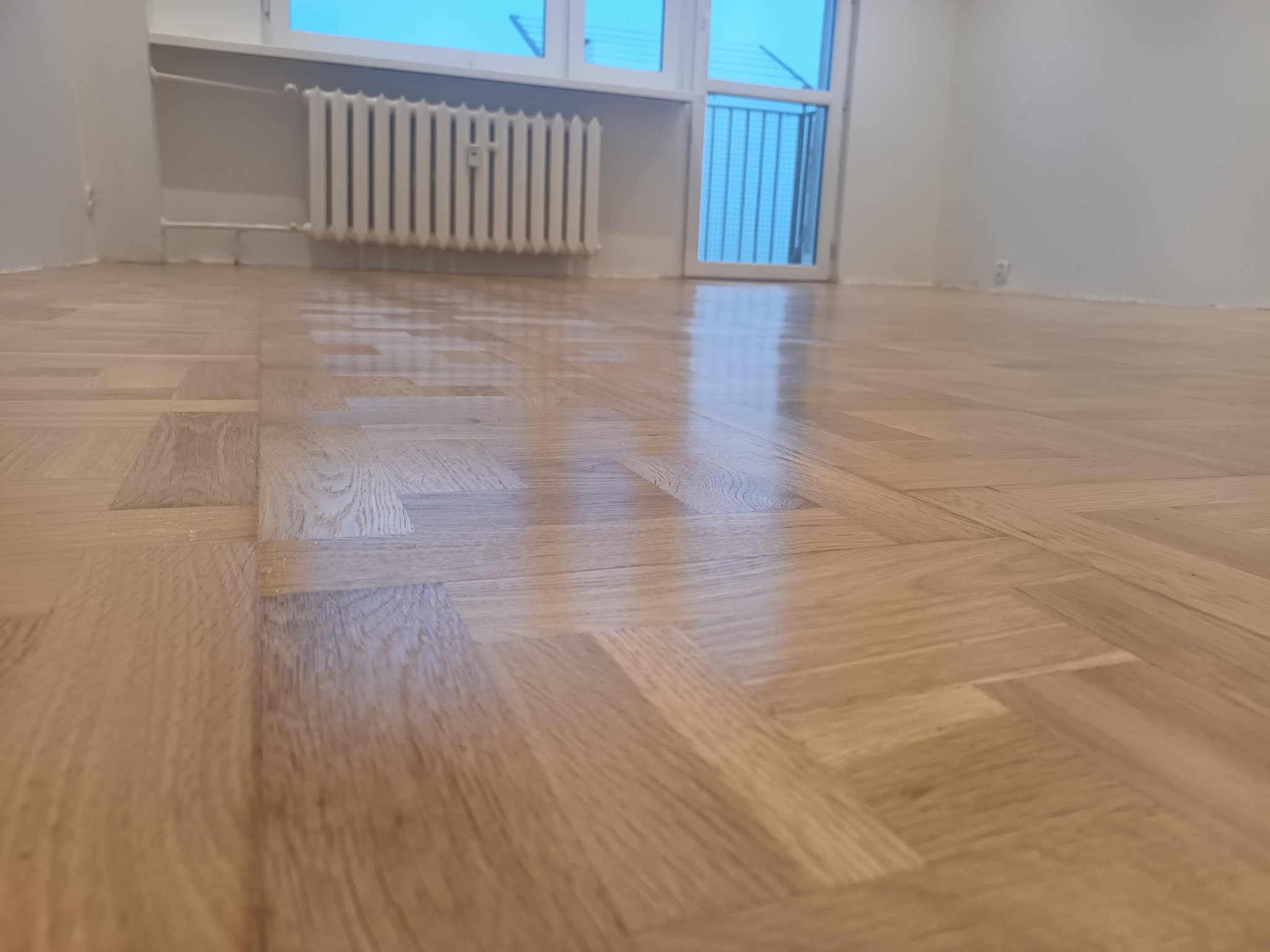 Świeżo położony parkiet ułożony w jodełkę, z widocznym kaloryferem i oknem w tle, sfotografowany z niskiej perspektywy.